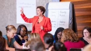 Peggy Vasquez in a red suit standing among a seated audience with her hand raised, teaching resilience for administrative professionals